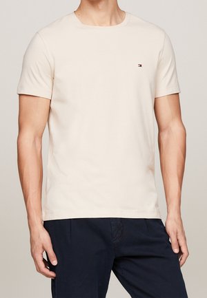 Man wearing a plain beige short-sleeve crew neck t-shirt with a small logo on the chest, paired with dark navy pants.