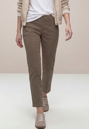 Persona con chaqueta beige, camisa blanca, pantalones marrones de corte recto y zapatillas beige caminando hacia adelante sobre un fondo liso.