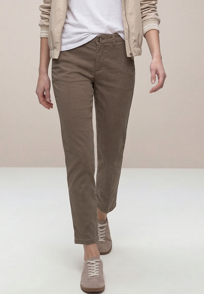 Persona con chaqueta beige, camisa blanca, pantalones marrones de corte recto y zapatillas beige caminando hacia adelante sobre un fondo liso.