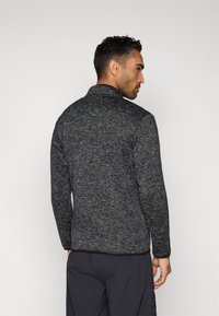 Μαύρο και γκρι μπουφάν από μαλακό φλις με ψηλό γιακά, μακριά μανίκια και ίσια ραφή στο τελείωμα, εμφανιζόμενο από πίσω. Υφασμάτινη υφή.