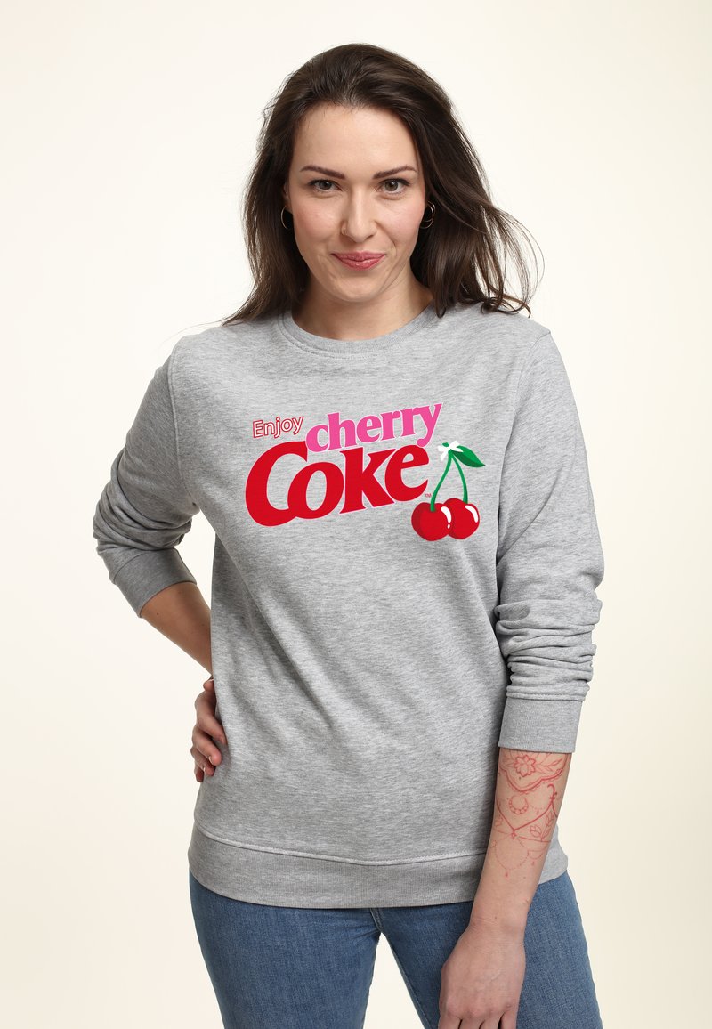 Felpa grigia chiaro con la scritta "Goditi la cherry Coke" in lettere rosse e rosa in grassetto, caratterizzata da due ciliegie rosse e foglie verdi. Polsini e orlo a costine.
