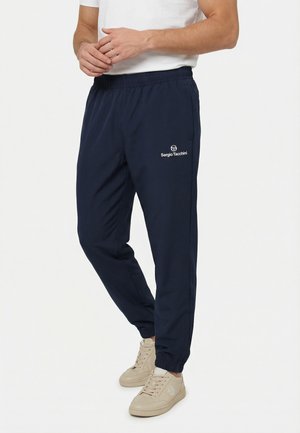 Hombre con pantalones deportivos azul marino de Sergio Tacchini, camiseta blanca y zapatillas beige, de pie con las manos entrelazadas.