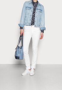 Giacca di jeans azzurro chiaro sopra una camicia a motivi blu navy, jeans skinny bianchi, sneakers bianche e una borsa a secchiello in pelle blu con accenti blu navy.