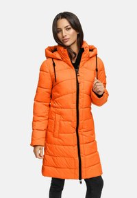 Mujer vestida con un abrigo acolchado largo de color naranja brillante, con capucha y cremallera negra, de pie frente a un fondo claro y sencillo.