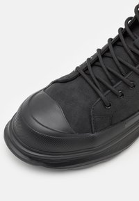 Černá sportovní bota s nubukovým svrškem, gumovou špičkou, texturovanou podrážkou a kulatými tkaničkami, s kovovými oky pro šněrování.