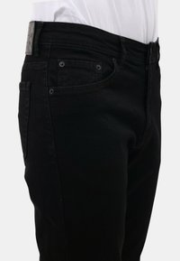 Man in zwarte spijkerbroek met zichtbare voor- en zijzakken, gecombineerd met een ingestopt wit shirt, afgebeeld van de taille tot halverwege de dijen.