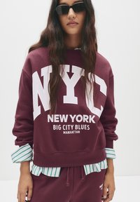 Felpa bordeaux con grande testo bianco "NYC", grafica "NEW YORK" e "MANHATTAN". Indossata sopra una camicia a righe verdi e bianche.