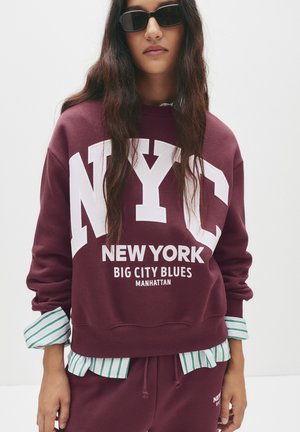 Felpa bordeaux con grande testo bianco "NYC", grafica "NEW YORK" e "MANHATTAN". Indossata sopra una camicia a righe verdi e bianche.