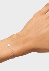 Pulsera de cadena de plata con pequeñas perlas blancas y un charm en forma de oso, llevada en una mano de tono de piel claro.