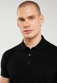 Mann mit kurzen Haaren und blauen Augen, der ein schwarzes Poloshirt trägt und vor einem einfarbigen hellen Hintergrund nach vorne schaut.
