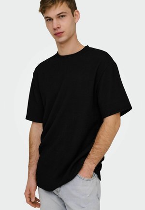 Jeune homme à la peau claire et aux cheveux courts portant une chemise noire ample à manches courtes et un pantalon gris clair, une main dans la poche.