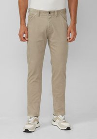 Man in beige casual pants en witte sneakers, staand tegen een effen grijze achtergrond.
