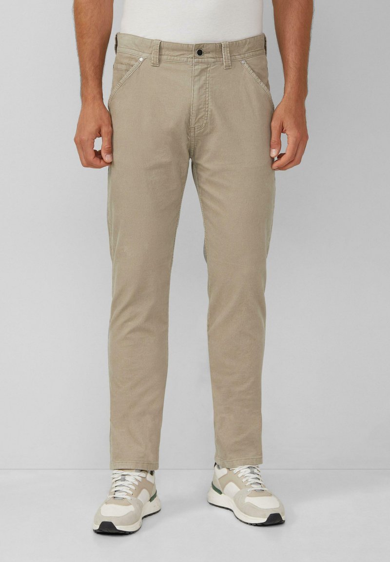 Man in beige casual pants en witte sneakers, staand tegen een effen grijze achtergrond.