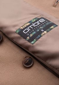 Beige Jacke aus Stoff mit strukturierter Oberfläche, ausgestattet mit einem schwarzen Etikett mit grünen und gelben Akzenten sowie einem braunen Knopf zum Verschließen.