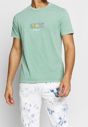 Miętowa bawełniana koszulka z okrągłym dekoltem z przodu ozdobiona jest wielokolorowym nadrukiem z logo Levi's w formie prostokąta. Noszona z białymi spodniami w stylu tie-dye.