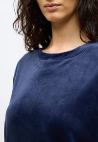 Γυναίκα με σγουρά σκο ύρα μαλλιά που φοράει μαλακό, βελούδινο μπλε ναυτικό τοπ με στρογγυλή λαιμόκοψη, εμφανίζεται από το πηγούνι έως το στήθος σε απλό φόντο.