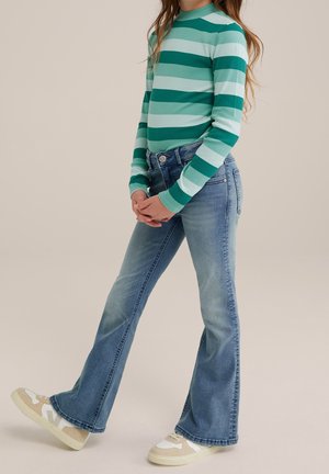 Enfant portant un t-shirt à manches longues rayé turquoise et bleu clair, un jean évasé bleu et des baskets beige et blanches, debout, mains jointes.