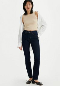 Mörkblå, figursydda jeans med hög midja, matchade med en beige ärmlös topp och en vit skjorta med knäppning. Enkla svarta ballerina-skor fulländar looken.