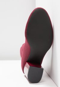 Bottine chaussette tricotée rouge avec semelle noire lisse et talon bloc, positionnée semelle vers le haut contre un mur et un sol blancs.