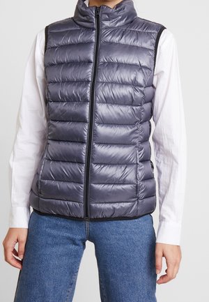 Personne portant une doudoune matelassée sans manches gris foncé zippée sur une chemise blanche à manches longues et un jean bleu sur un fond clair.