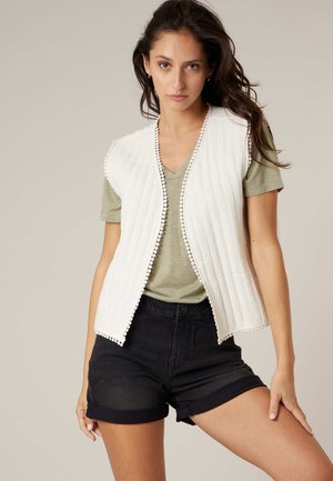 Jeune femme aux longs cheveux noirs porte un gilet matelassé blanc sans manches par-dessus un t-shirt vert à col en V et un short noir retroussé, posant avec la main sur la cuisse.