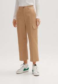Pantalons cargo beiges avec une coupe droite, dotés de poches latérales et d'un fermeture à bouton. Portés avec des baskets blanches et un haut décontracté.