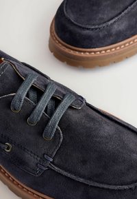 Pantofii din suede albastru închis au un vârf rotund, cusături în contrast și ocluri metalice cu șireturi bleumarin. Talpa din cauciuc are o finisare texturată.