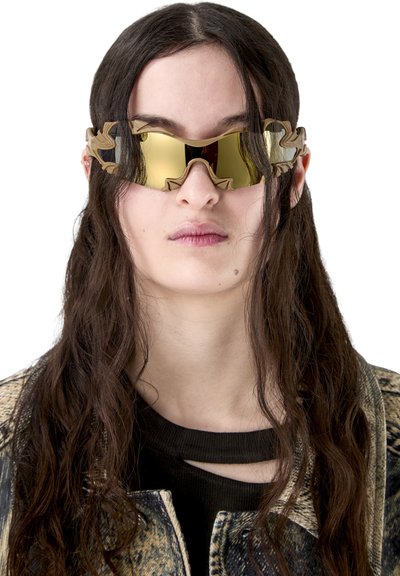 Jeune femme aux longs cheveux bruns portant des lunettes de soleil futuristes dorées réfléchissantes et une veste à motifs par-dessus un haut noir à décolleté découpé.