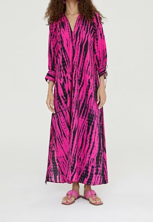 Kobieta ubrana w długą sukienkę z efektem tie-dye w kolorach różowym i czarnym, z rękawami do łokcia i dopasowanymi różowymi sandałami, na jednolitym tle.