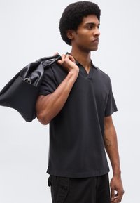 Νέος άνδρας με αφροκάρυ hairstyle φοράει μαύρο πουκάμισο και παντελόνι, κρατώντας μια μαύρη τσάντα στον ώμο του, κοιτάζοντας προς τα πλάγια μπροστά σε απλό φόντο.