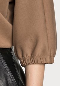 Braunes Langarmshirt mit elastischen Bündchen an den Armen, neben schwarzen Lederhosen, auf Ellbogenhöhe gekürzt, neutraler Hintergrund.