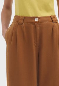 Pantalons plissés marron avec une texture lisse, dotés de poches latérales et d'un bouton argenté à la taille. Portés avec un top jaune.