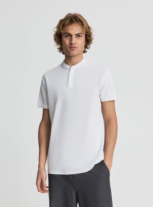 Giovane uomo con capelli biondi ricci che indossa una maglietta henley bianca a maniche corte e pantaloni scuri, in piedi davanti a uno sfondo semplice.