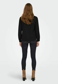 Blusa nera a maniche lunghe abbinata a jeans scuri aderenti. La modella indossa stivali alla caviglia in suede beige con tacco spesso. I capelli sono mossi.