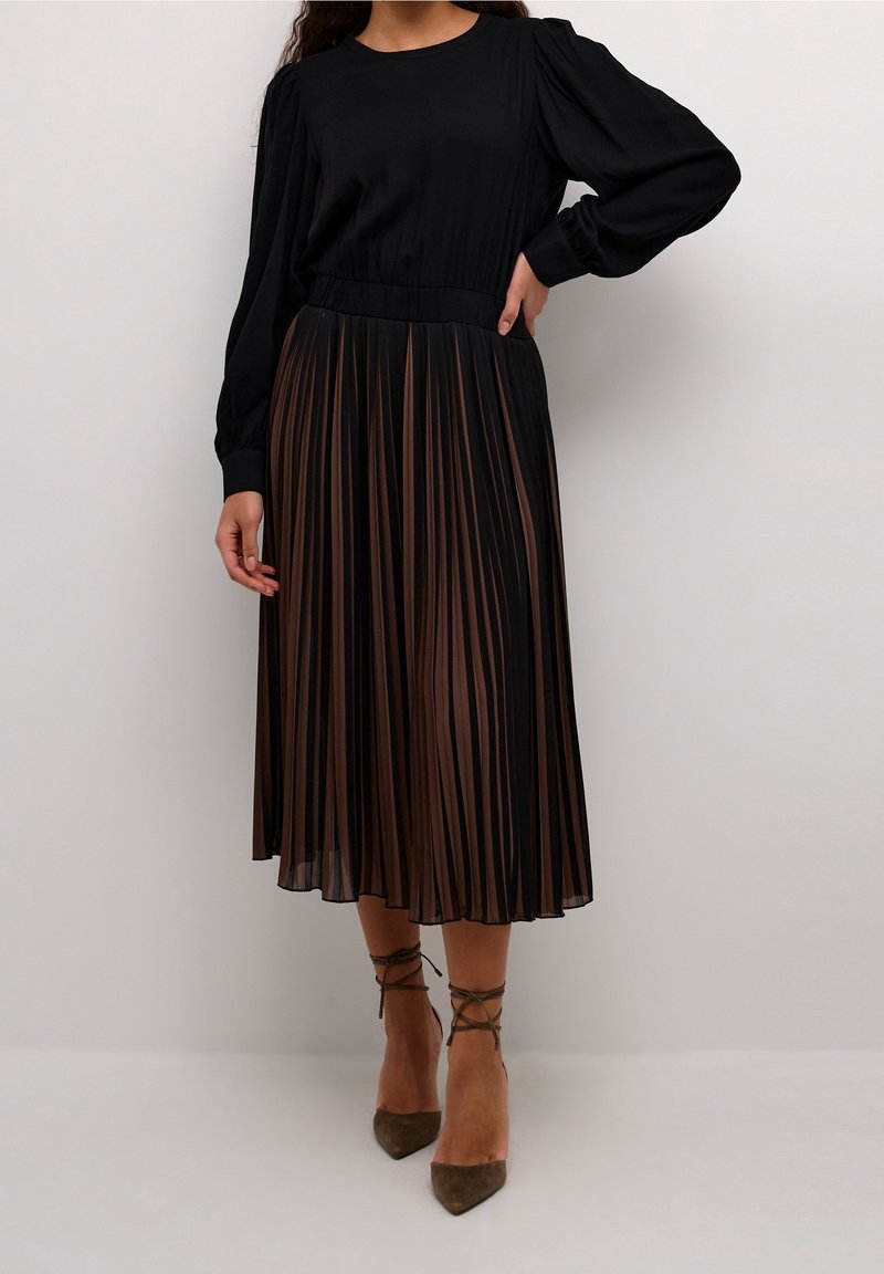 Haut noir à manches longues avec une jupe midi plissée marron. La jupe présente des rayures verticales et une taille ajustée. Le mannequin porte des talons marron.