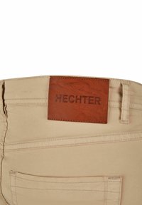 Beige broek met een gladde textuur, voorzien van een bruine leren label met "HECHTER" in reliëf op de taille en klassieke stikdetails.