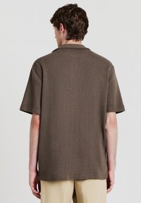 Camicia marrone a maniche corte con tessuto strutturato, vestibilità ampia e colletto piatto. Costruzione senza cuciture e orlo uniforme.