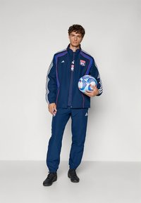 Marineblå atletisk joggedress med hvite striper, merkevarelogo på jakken, tettsittende bukser, holder en mønstret fotball. Mørke sko.