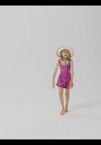 Niña joven vistiendo un mono rosa con estampados de frutas y un sombrero de ala ancha, caminando descalza sobre un fondo blanco liso.