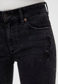 Vaqueros de mezclilla negros con una cintura alta, diseño de cinco bolsillos y detalles desgastados con bordes deshilachados cerca del muslo derecho.