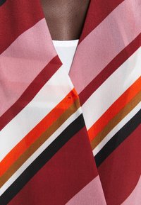 Gros plan sur une poitrine à peau foncée portant un vêtement rayé multicolore aux tons rouges, roses, blancs, orange, marron et noir.