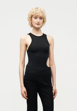 Ξανθός με σγουρά μαλλιά που φοράει μαύρο αμάνικο τοπ με πλαϊνή κοπή και μαύρο παντελόνι, στέκεται μπροστά σε ένα απλό φωτεινό φόντο.