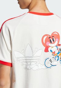 Krém színű póló piros kiegészítőkkel, amelyen egy kerékpáron ülő mesefigura és a hátsó oldalon egy grafikus Adidas logó található. Pamut anyag.