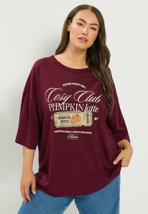 Donna che indossa una camicia larga color bordeaux con testo "Cozy Club Pumpkin Latte" e grafica di un biglietto per il campo di zucche, abbinata a jeans blu.