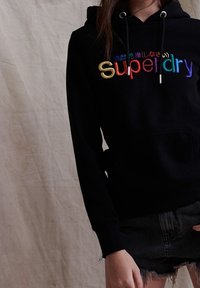 Černá mikina s kapucí a šňůrkami, která má multibarevné vyšívané logo "Superdry". Přiléhavý design a jemný materiál, zkombinováno s tmavými šortkami.