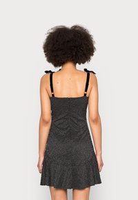 Schwarzes Polka-Dot-Kleid mit dünnen Trägern, ausgestattet mit einem fransigen Saum und Bindebändern an den Trägern. Glattes Gewebe mit leichtem Stretch.