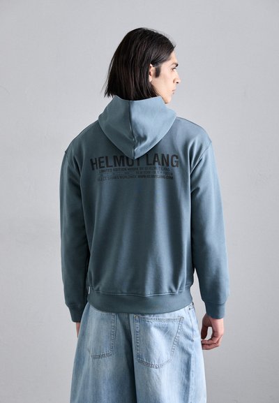 Veste à capuche en polaire bleu-gris avec un cordon de serrage. Le dos présente un texte de logo noir et l'impression "ÉDITION LIMITÉE". Style ample, associée à un short en denim clair.