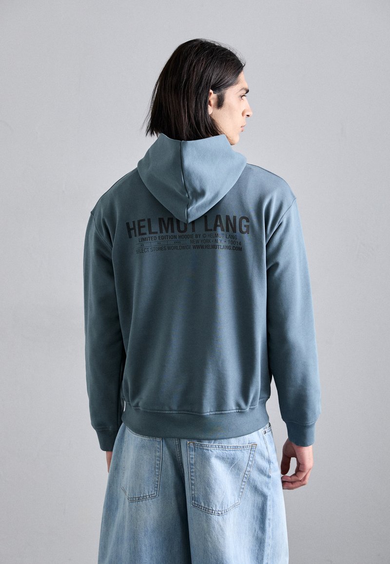 Hanorac din fleece gri-albastru cu glugă ajustabilă. Partea din spate prezintă un text logo negru și inscripția „EDIȚIE LIMITATĂ”. Stil lejer, asortat cu pantaloni scurți din denim deschis.