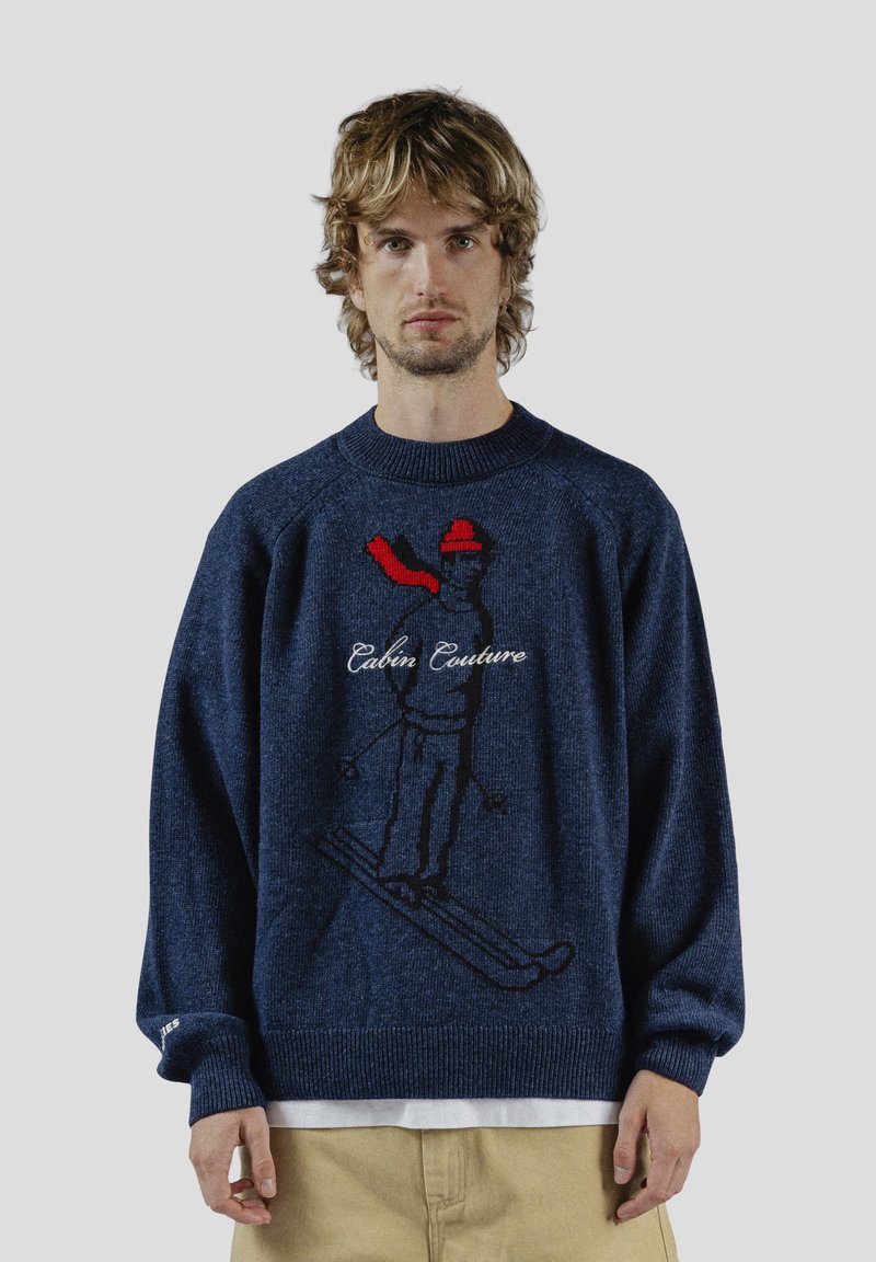 Laivastonsininen neulepaita, jossa on musta-punainen kirjailtu hiihtäjäkuvio ja alla teksti "Cabin Culture". Pehmeä pinta ja rento istuvuus.