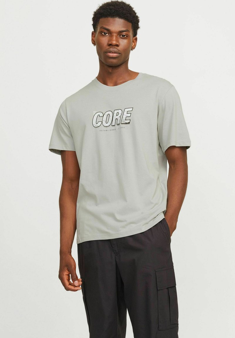 Mann in hellgrauem T-Shirt mit "CORE"-Aufdruck und schwarzen Cargo-Hosen, der mit einer Hand in der Tasche vor einem schlichten Hintergrund steht.